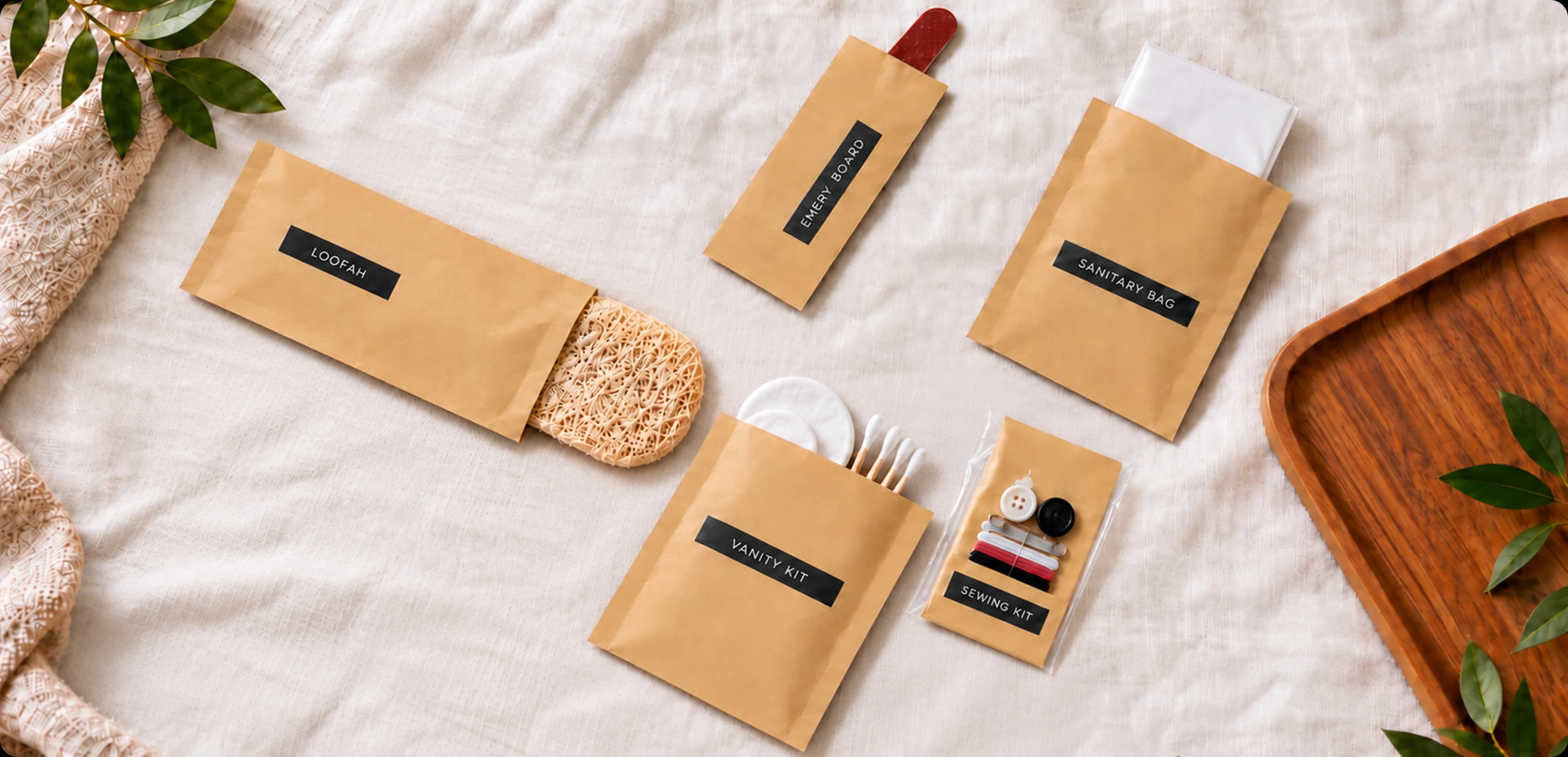 Kraft paper amenity pouches — loofah, emery board, sanitary bag, vanity kit and sewing kit — on white linen beside a wooden tray